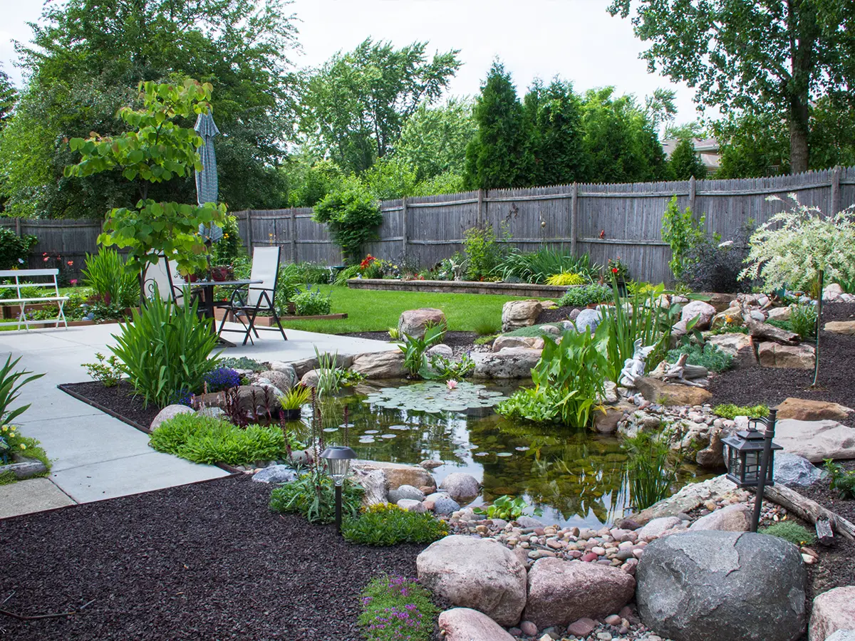 Backyard pond with natural stone border, lily pads, and surrounding greenery beside a modern patio seating area