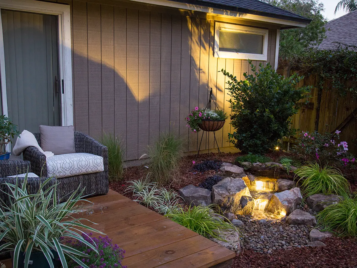 Cozy backyard seating area with illuminated garden waterfall feature, surrounded by plants and a wooden deck