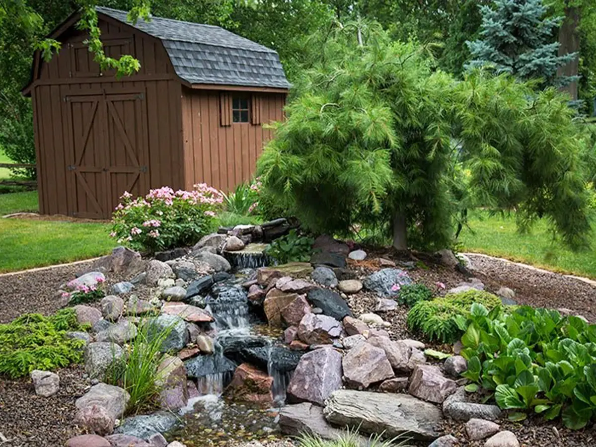 Rustic garden waterfall with natural stones and greenery, set beside a small wooden barn in a landscaped yard