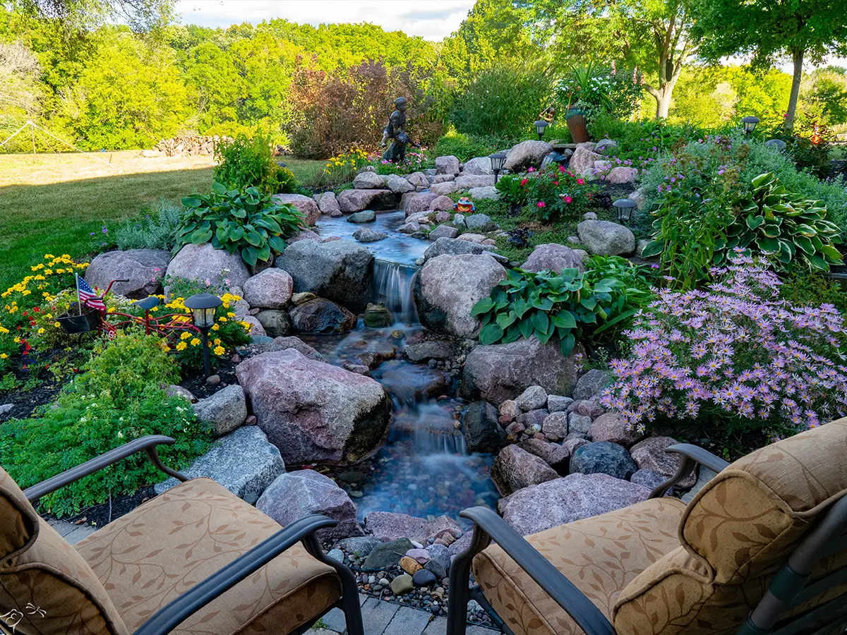 Backyard seating area overlooking a cascading waterfall garden with natural rocks, lush greenery, and vibrant flowers, creating a peaceful outdoor retreat