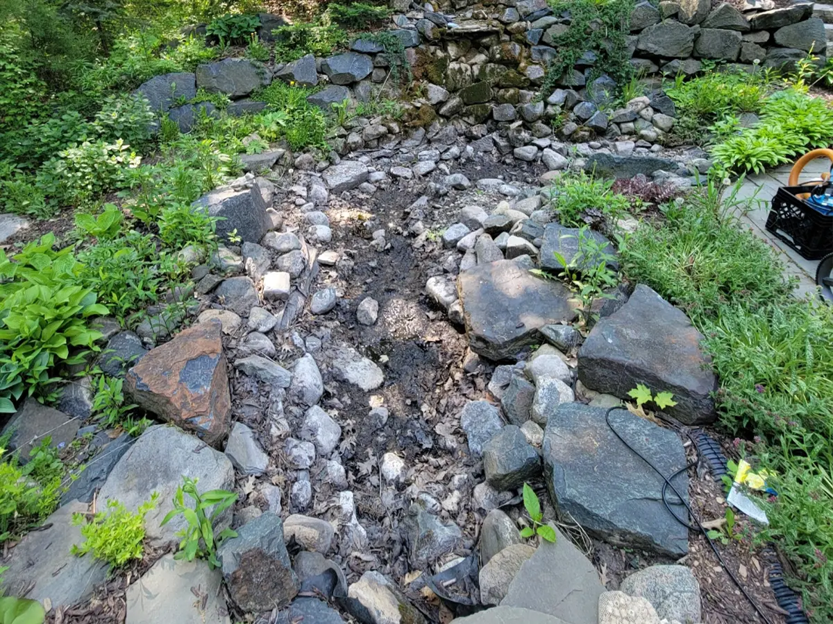 Unfinished garden stream with dry rocks and stone lining, showing initial layout in backyard landscape.