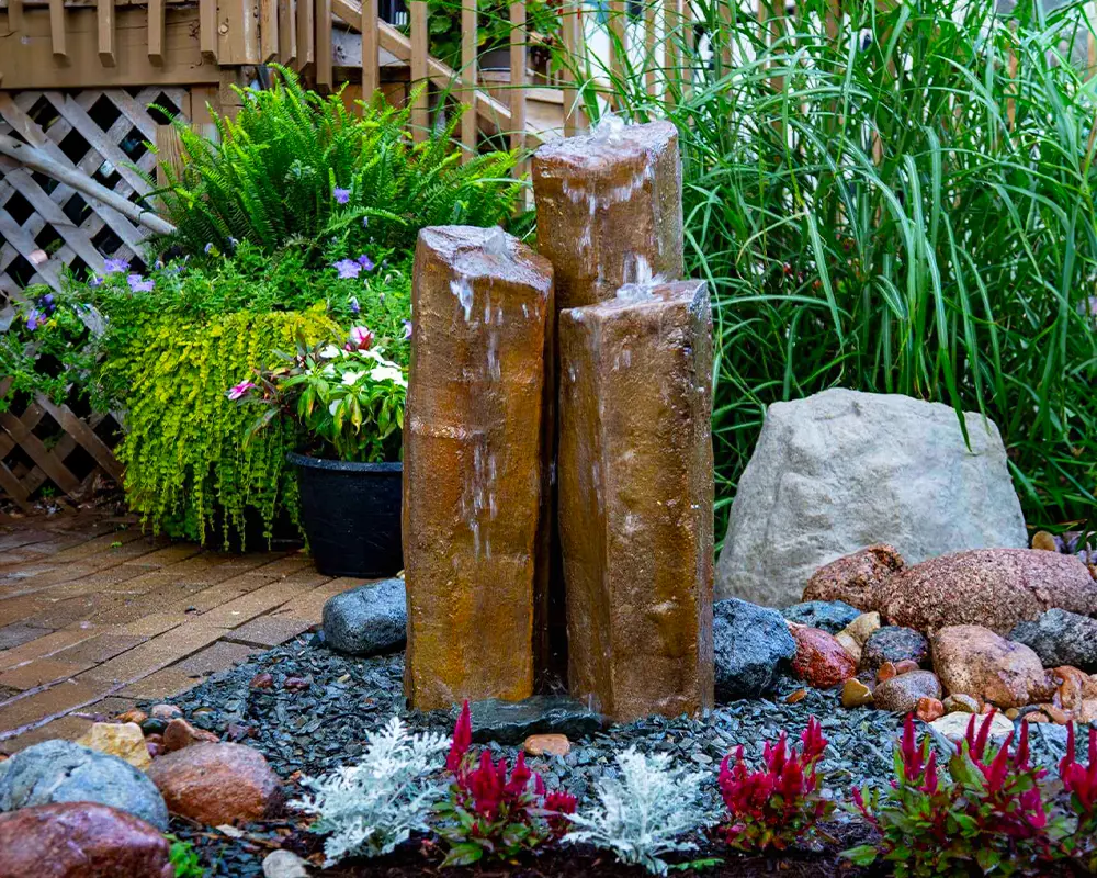 Triple column fountain with cascading water, surrounded by greenery and decorative rocks in a garden setting