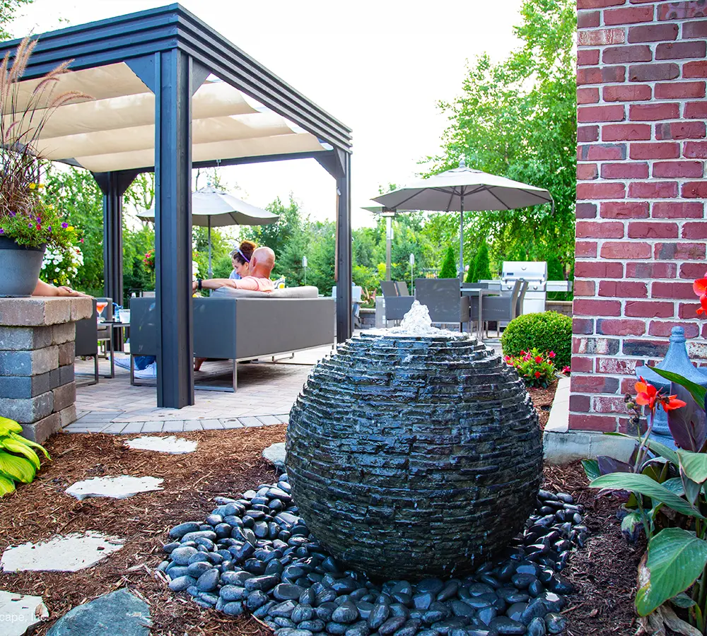 Round stacked-stone fountain with bubbling water, set on a pebble base near a cozy backyard patio