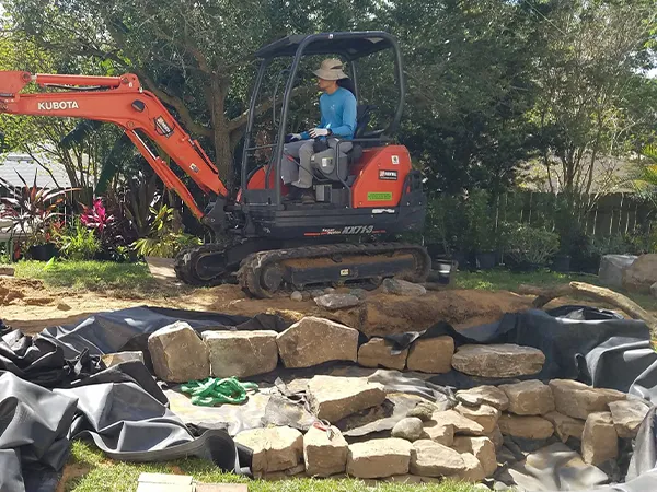 Excavator preparing site with rocks for new pond construction in a residential backyard