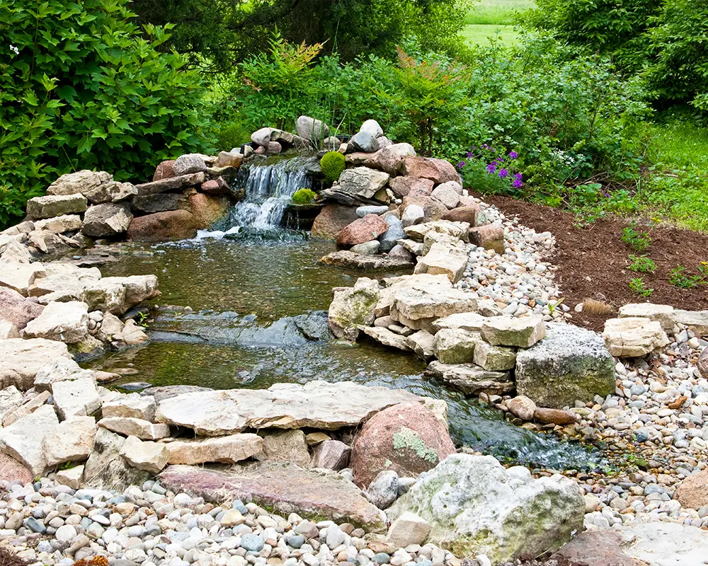 Backyard pond with waterfall feature under repair, surrounded by natural stone and greenery.