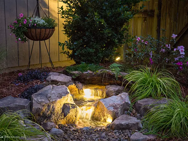 Illuminated pondless waterfall feature in a garden, surrounded by decorative rocks and plants.